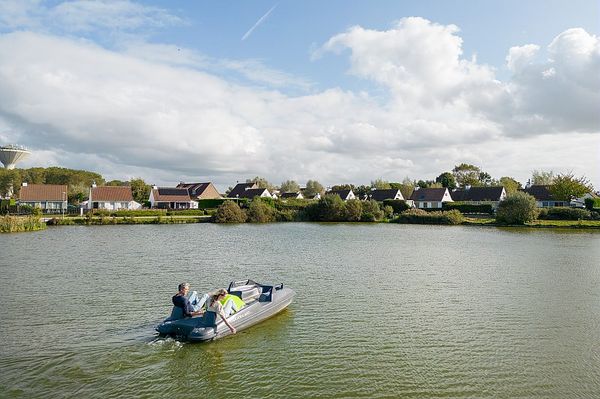 Un couple profite d'une balade en pédalo sur un lac, avec en arrière-plan des cottages entourés de verdure, sous un ciel partiellement nuageux.