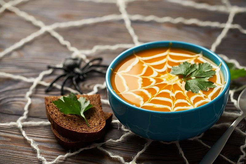 Bol de soupe orange décorée d’une toile d’araignée en crème, garni de persil et accompagné de tranches de pain, sur un thème Halloween.