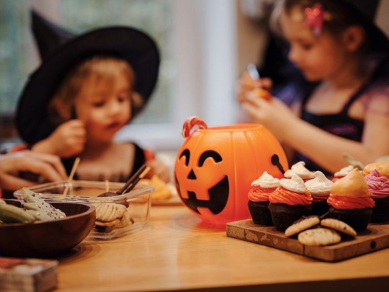 Enfants déguisés, assis autour d’une table décorée pour Halloween, avec un seau citrouille et des cupcakes colorés au premier plan.