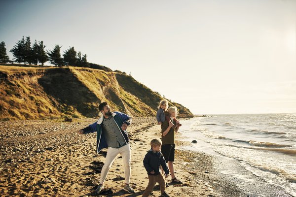 Une famille profite d’une balade sur la plage au coucher du soleil, un adulte lance des galets pendant que les enfants jouent au bord de l’eau.