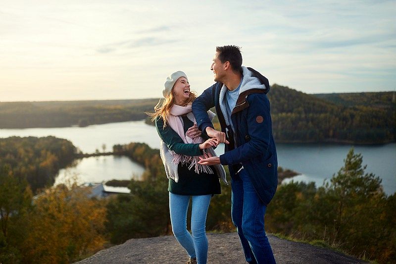 Un couple en tenue d’automne rit et se tient par les mains sur un point de vue, avec un lac et une forêt aux couleurs dorées en arrière-plan.
