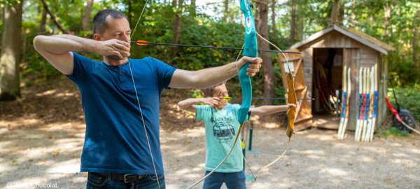 Un homme et un enfant pratiquent le tir à l'arc en pleine nature, concentrés et équipés de leurs arcs. À l'arrière, un cabanon en bois stocke du matériel supplémentaire.