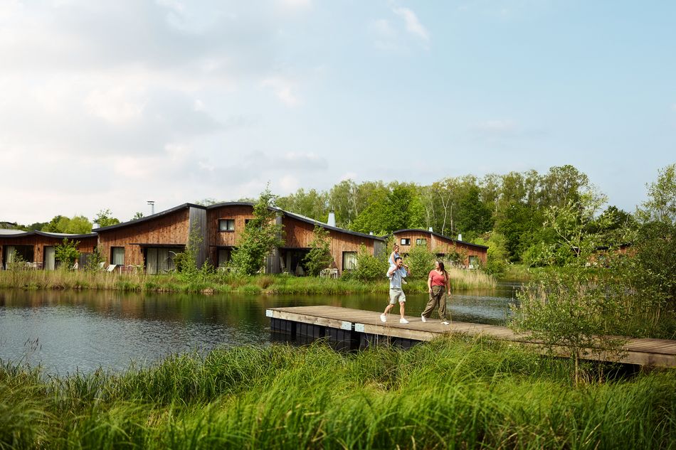 Un couple marche sur un ponton en bois au bord d'un étang, l'homme portant un enfant sur ses épaules. En arrière-plan, des cottages en bois entourés de verdure offrent une ambiance de villégiature en pleine nature.