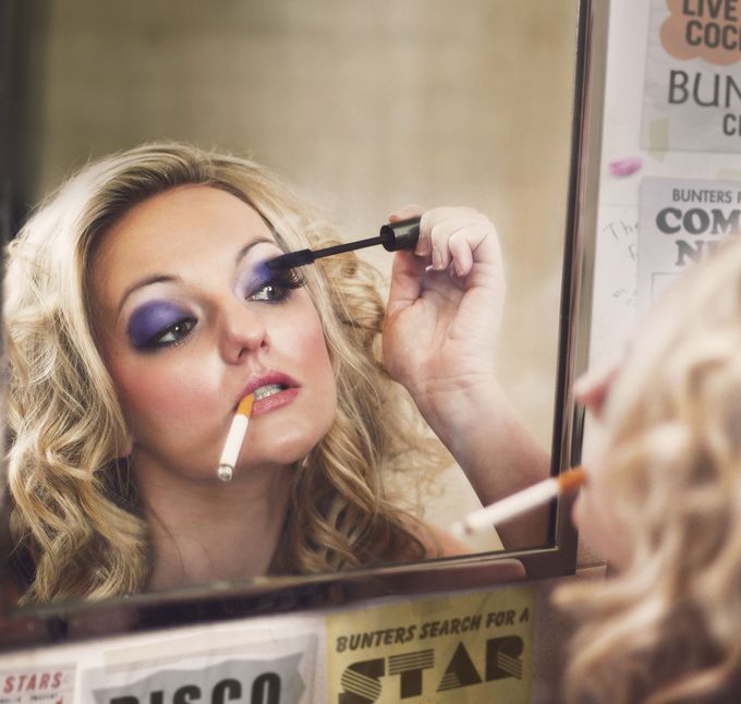 Blonde woman applying mascara in mirror whilst smoking a cigarette