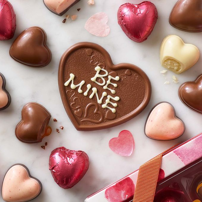 Chocolate heart with 'be mine' written with white icing, layed flat amongst smaller chocolate hearts