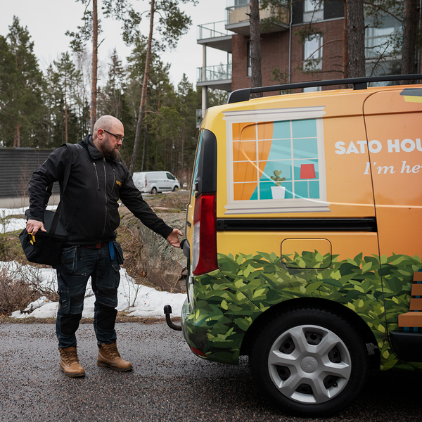 Talomestari Sami Korhonen tummassa asussa talon pihalla edessään talomestarin keltainen auto. Maassa on lunta ja taustalla korkeita mäntyjä.
