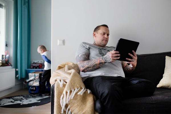 SATOkodin asukkaat Janne ja Joona kotosalla. Janne lukee sohvalla tablettia, Joona leikkii.